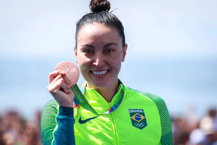 Mulher sorridente com medalha de bronze Rio 2016, jaqueta verde fluorescente.