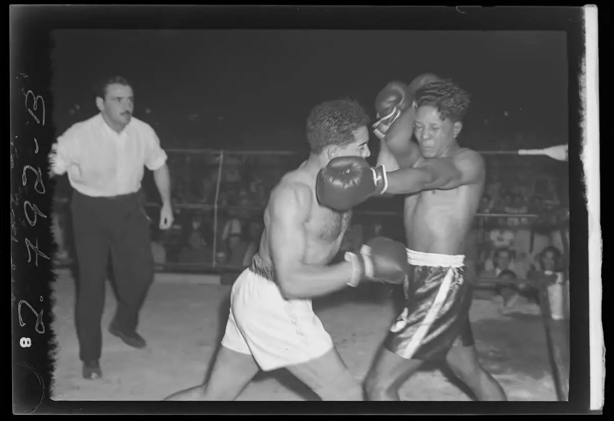 Luta de boxe em preto e branco, com árbitro e lutadores em ação.