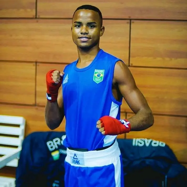 Boxeador em treino, uniforme azul, bandagens vermelhas, parede de madeira ao fundo.