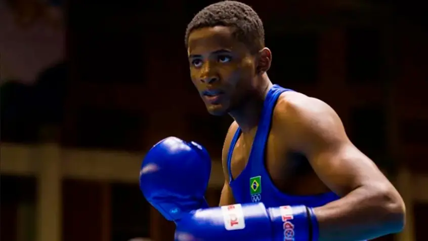 Boxeador em ação no ringue, uniforme azul com símbolo do Brasil e logotipo dos Jogos Olímpicos.