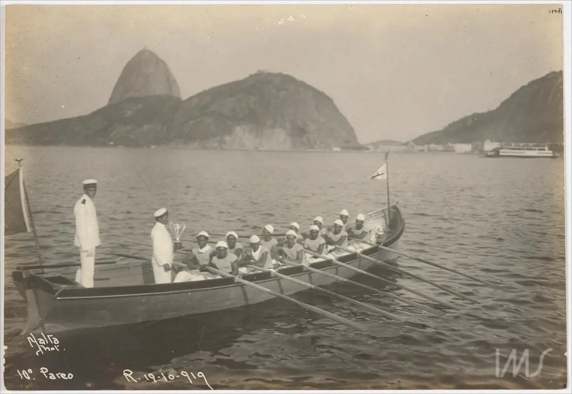 Grupo de remadores em barco, montanhas rochosas ao fundo.