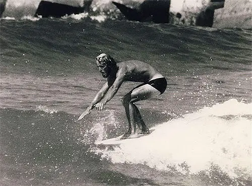 Surfista equilibrado em onda, praia ao fundo.