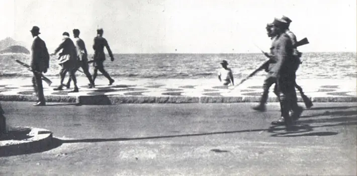 Homem com rifle caminha à esquerda; soldados avançam; homem olha para praia; soldados avançam pela calçada; praia e ondas ao fundo.
