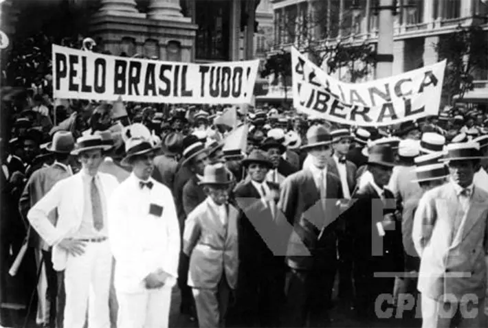 Multidão formal em comício, cartazes "PELO BRASIL TUDO!" e "ALIANÇA LIBERAL" em praça urbana.