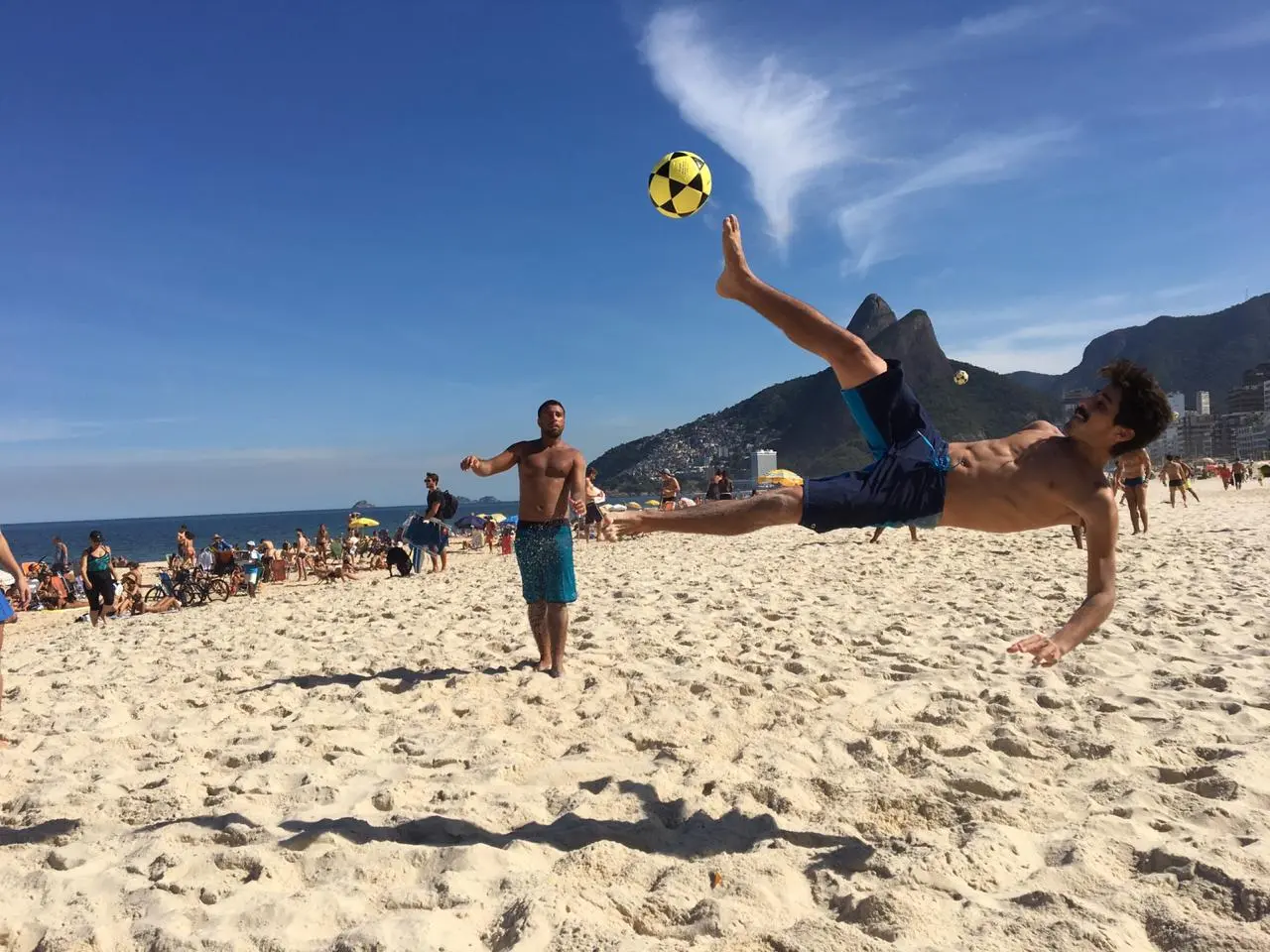Pessoas desfrutando da praia com futebol ao fundo.