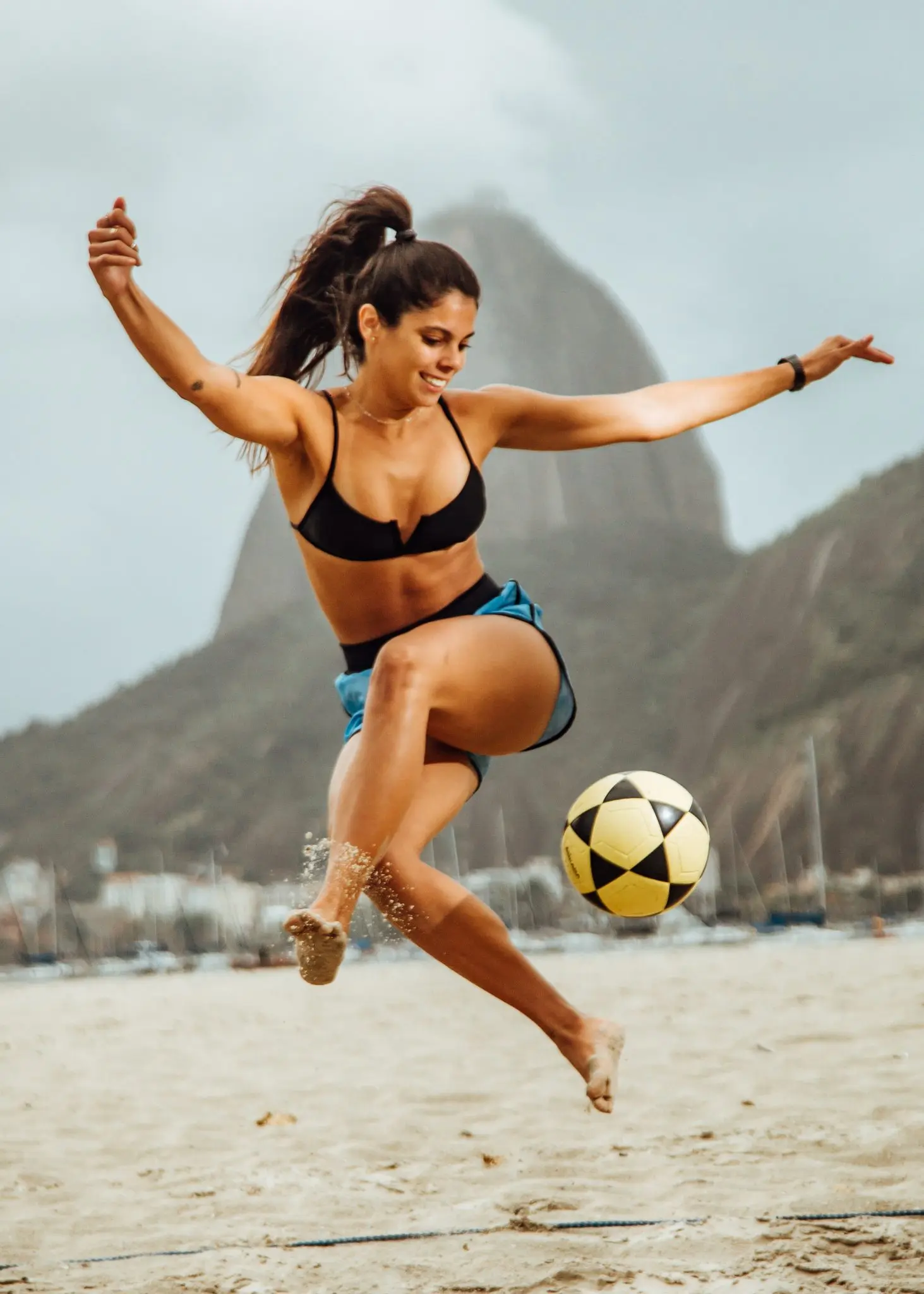 Mulher jogando futebol na praia, em movimento, com montanha ao fundo e céu nublado.