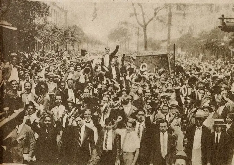 Multidão em rua, homem em plataforma, discurso ou celebração.