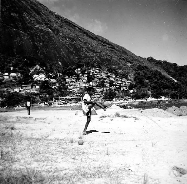 Homem jogando futebol em campo, com colina e casas ao fundo.