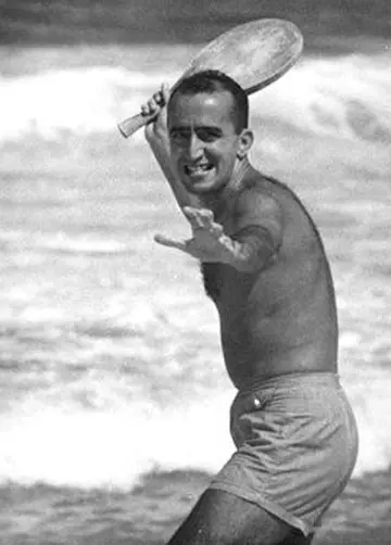 Homem sorridente segurando raquete de frescobol, ondas do mar ao fundo.