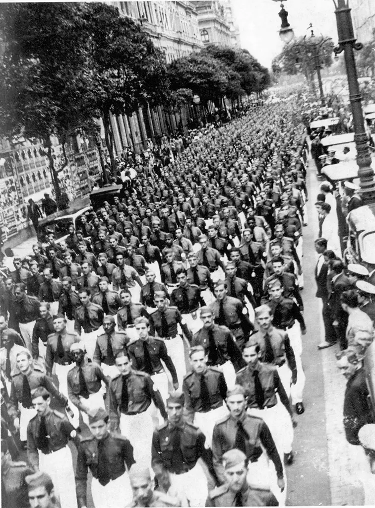 Desfile militar em formação, multidão uniformizada, árvores e construções antigas ao fundo.