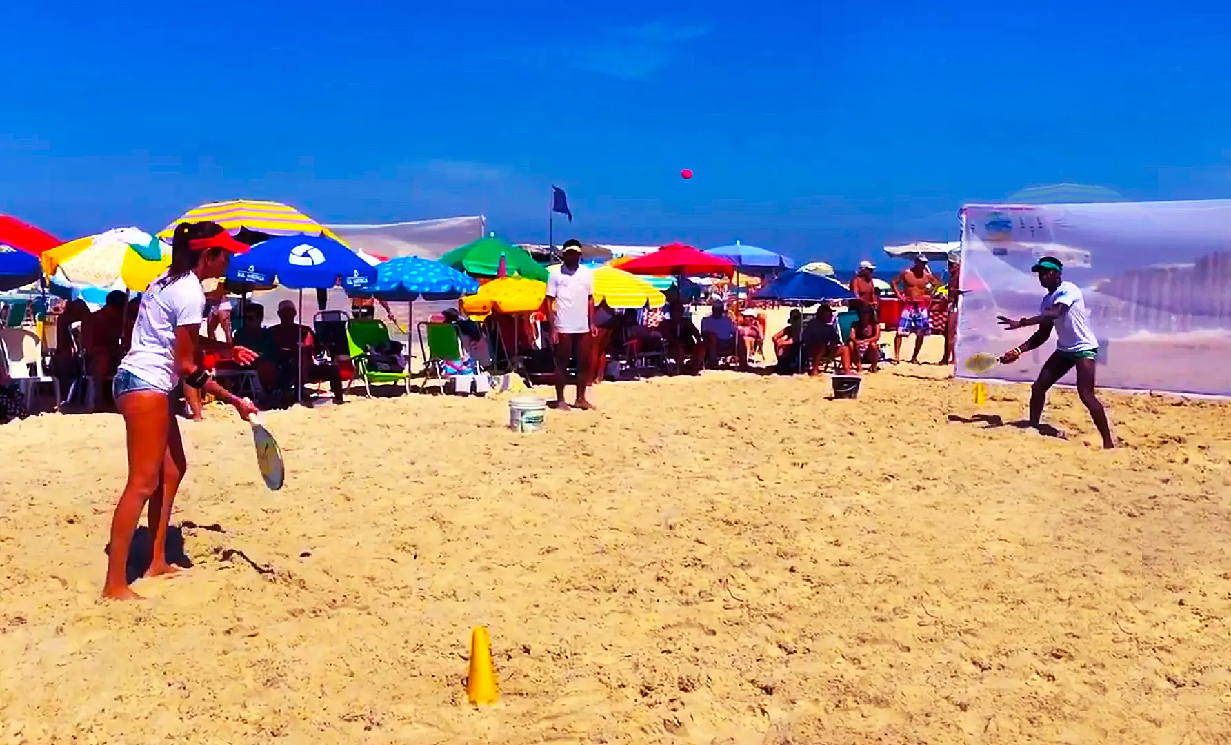 Praia lotada com pessoas jogando frescobol e relaxando sob guarda-sóis coloridos.