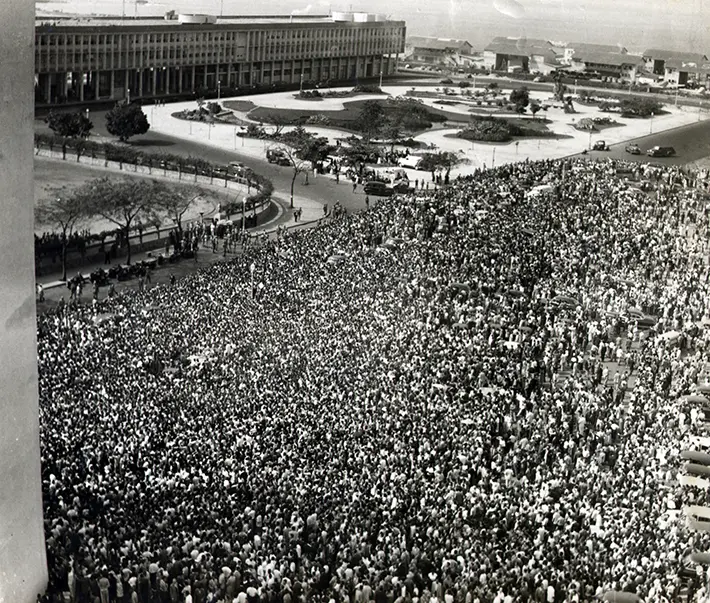 Multidão em parque com edifícios governamentais ao fundo.