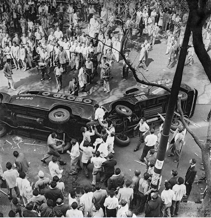 Cenário de motim com carros derrubados e multidão observando.