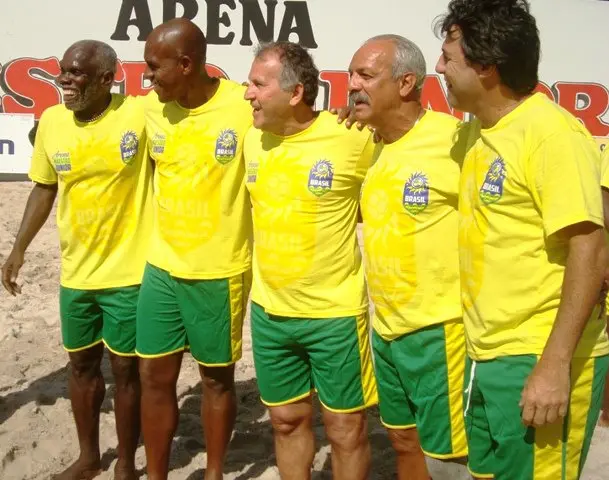 Cinco homens em uniforme amarelo com detalhes verdes e azuis sorriem para a câmera.