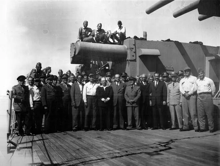 Grupo de pessoas em torno de canhão naval, uniformes militares e ternos formais.