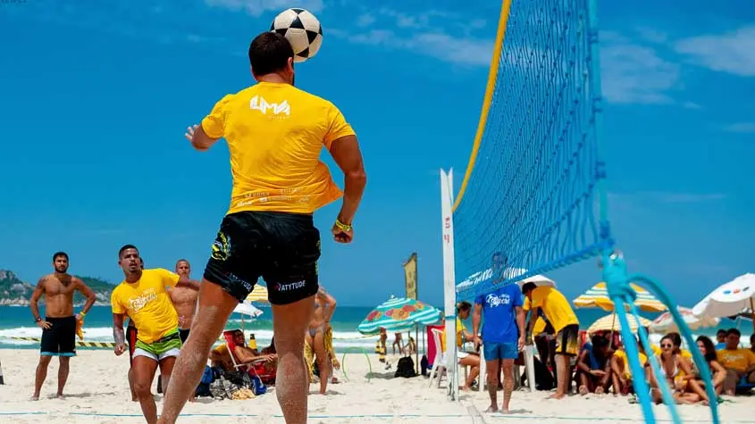 Praia com pessoas jogando futevôlei, céu azul e guarda-sóis coloridos.