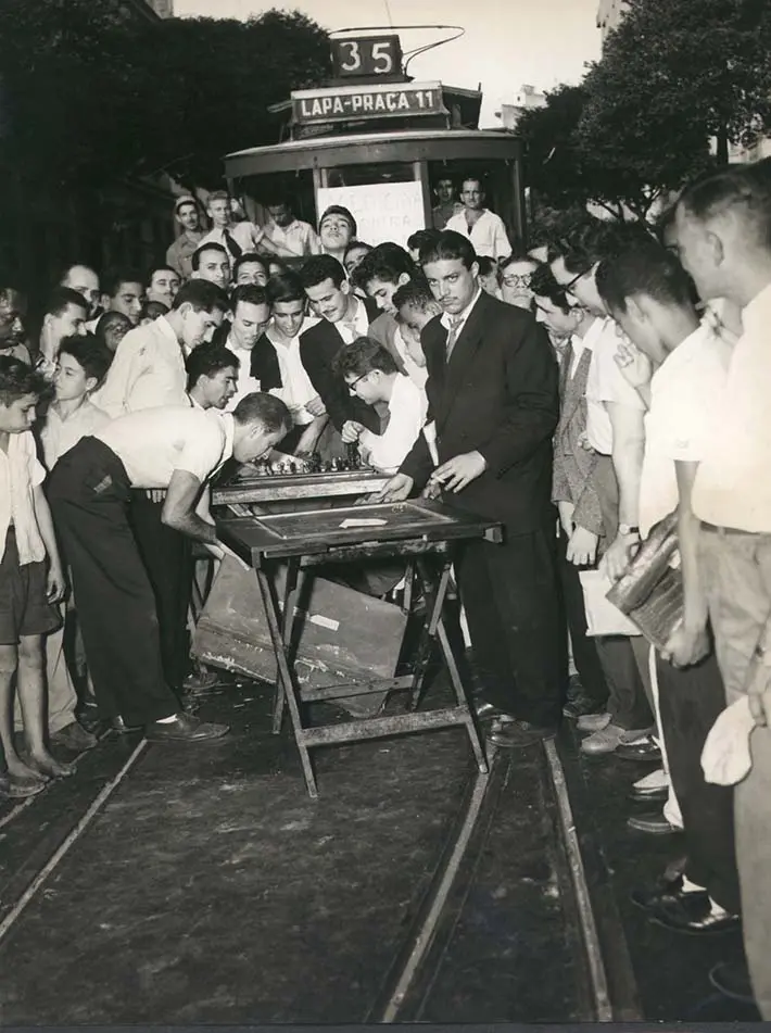 Evento público com pessoas observando objetos em mesa, bonde ao fundo.