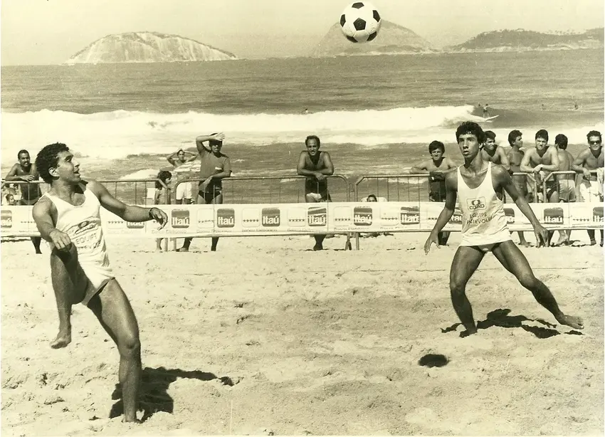 Jogador de futevôlei em ação na praia, com espectadores ao fundo.
