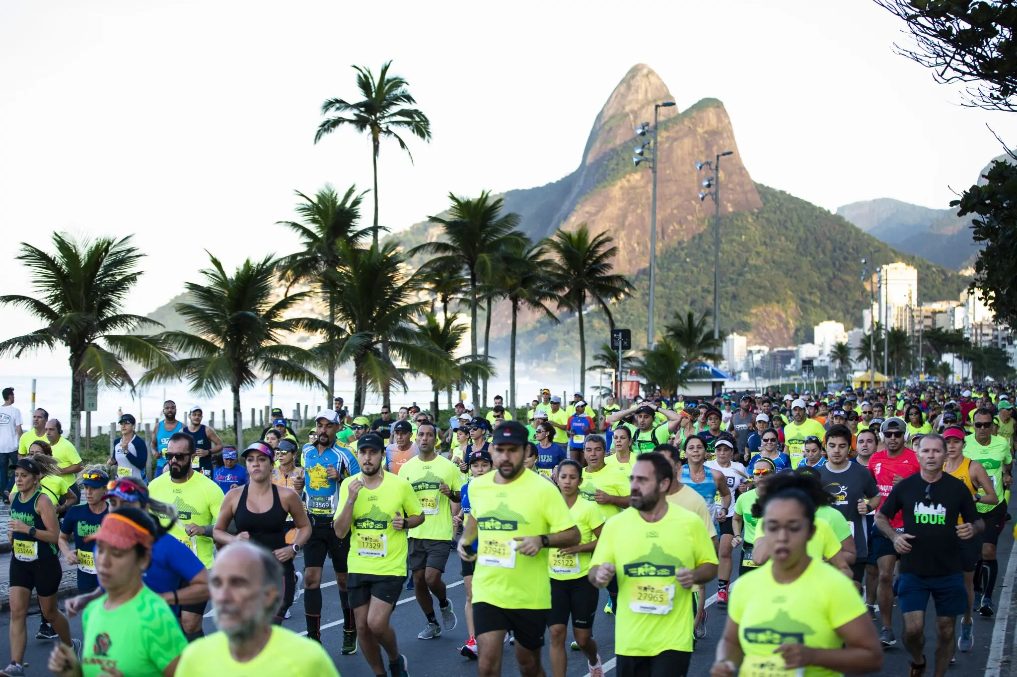 Corrida em praça costeira com palmeiras, mar calmo, corredores em fileiras, montanha rochosa e edifícios modernos ao fundo.