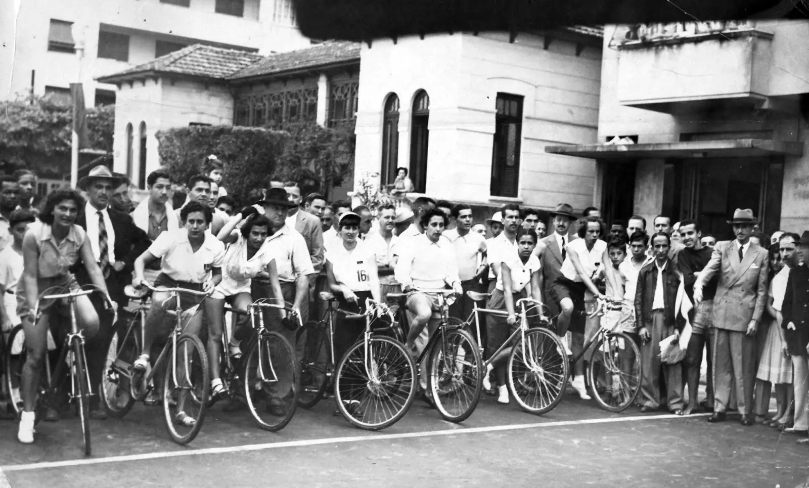 Grupo de pessoas em frente a edifício, bicicletas à esquerda, ternos à direita.