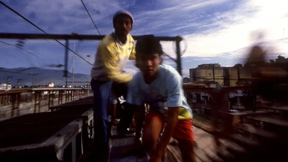 Dois jovens em um trem em movimento, urbano e montanhoso.