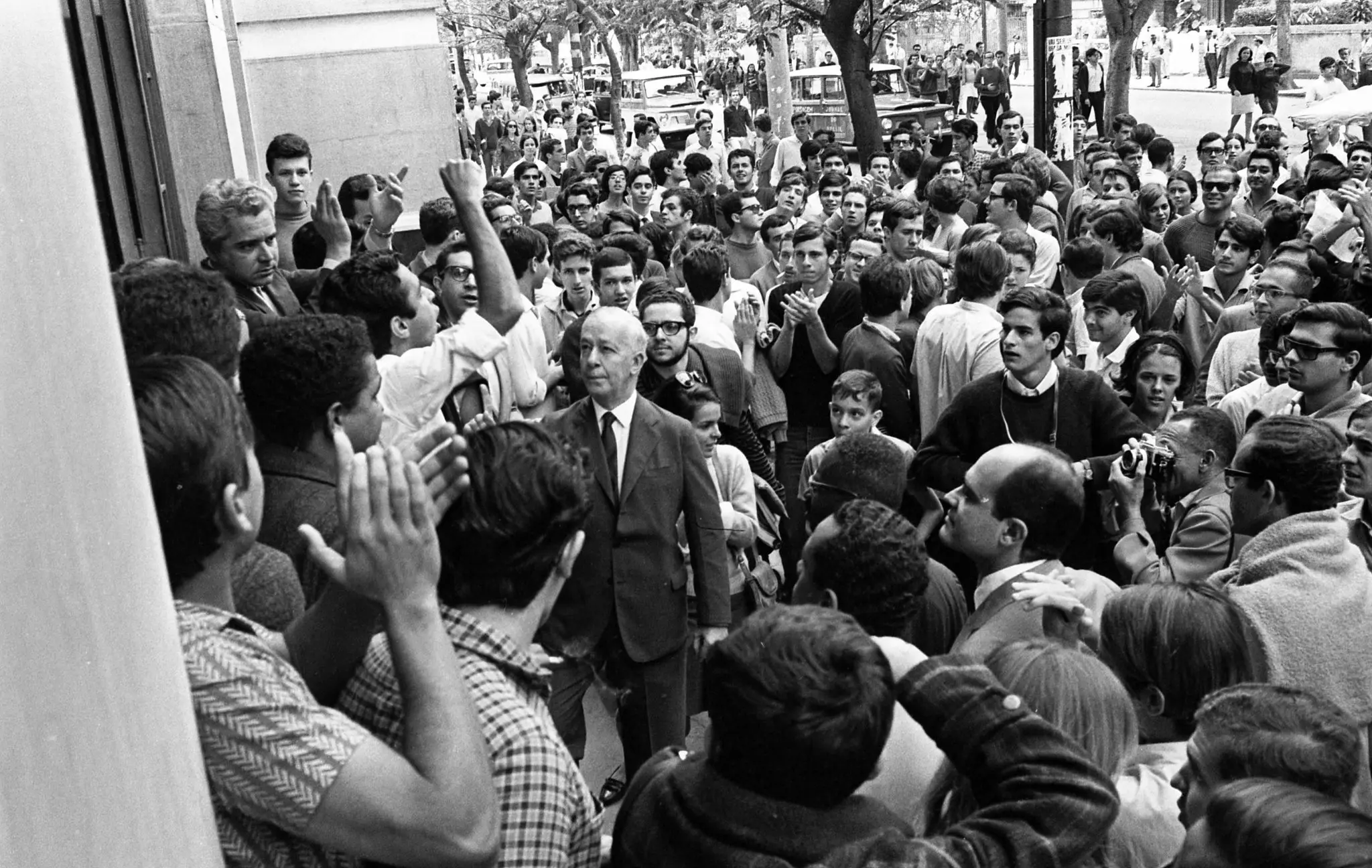 Aglomeração urbana em protesto, com destaque para homem em terno e estudantes erguendo mãos.