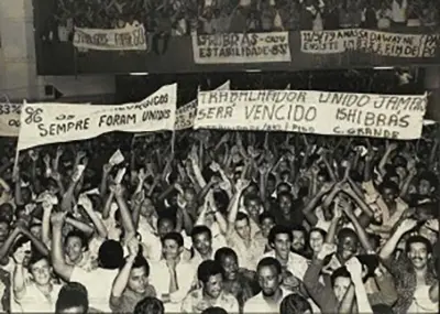 Multidão animada ergue bandeiras e levanta braços, frente para frente.