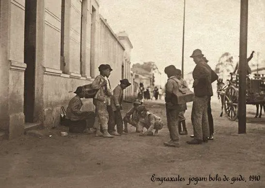 Grupo de pessoas em rua antiga, preto e branco, com edifício e atividades sociais.
