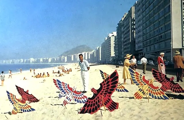 Praia animada com pessoas desfrutando do sol, mar calmo e edifícios ao fundo.
