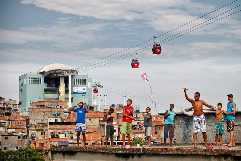 Um grupo de pessoas em telhado com pipas, edifícios e teleféricos ao fundo.