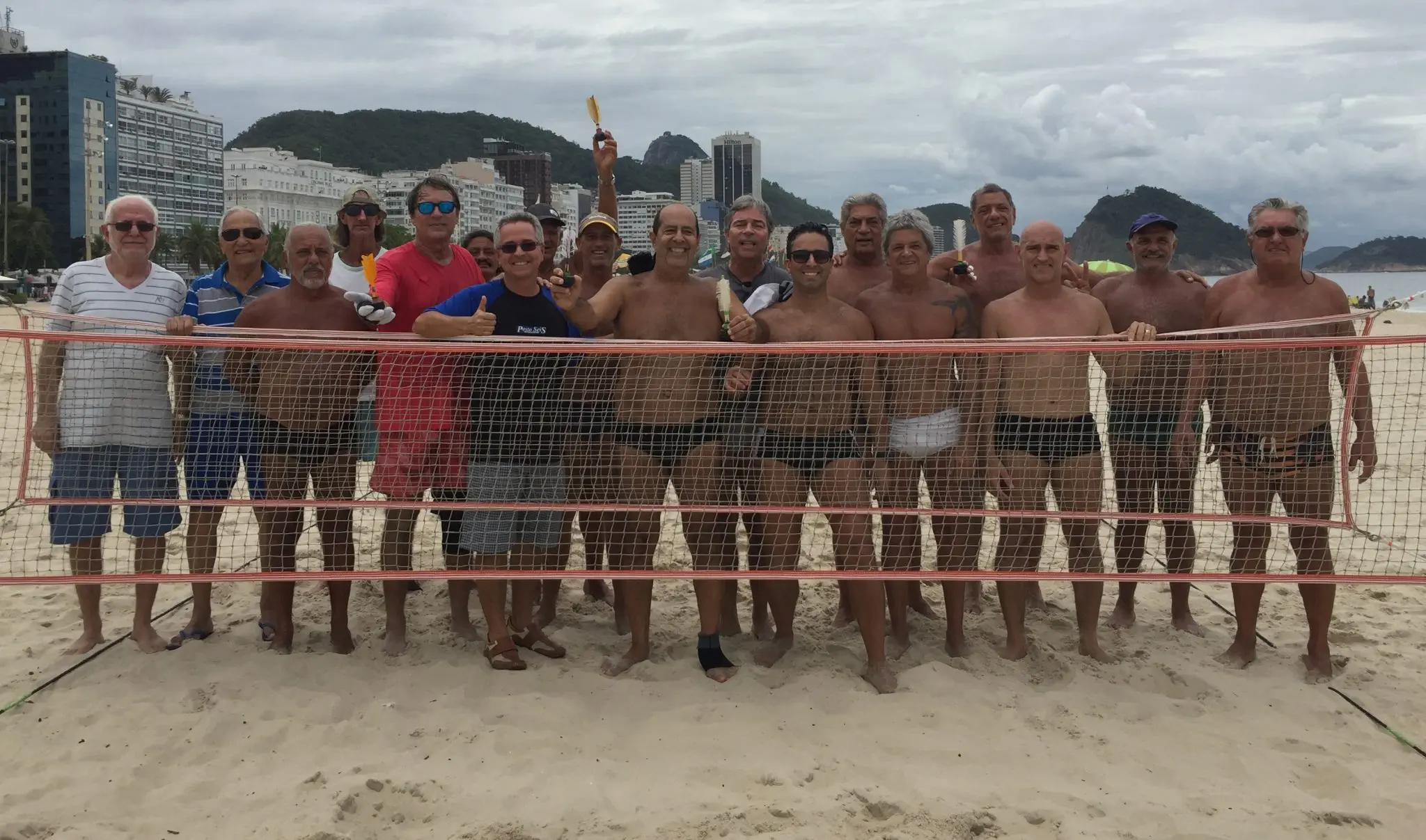 Grupo de homens em praia com rede de peteca, edifícios e montanha ao fundo.