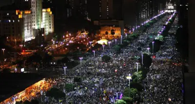 Multidão em protesto noturno, área urbana densa, edifícios altos e monumento iluminado.