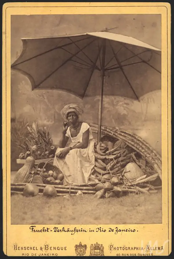 Mulher sentada em banco de bambu, protegida por guarda-sol, com frutas e vegetais ao redor.