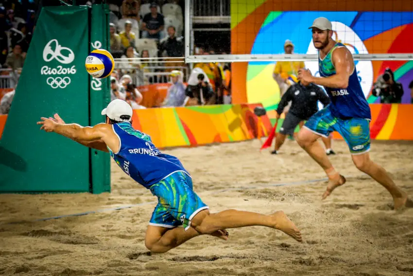 Jogador azul salta para bloquear a bola em jogo de vôlei de praia.