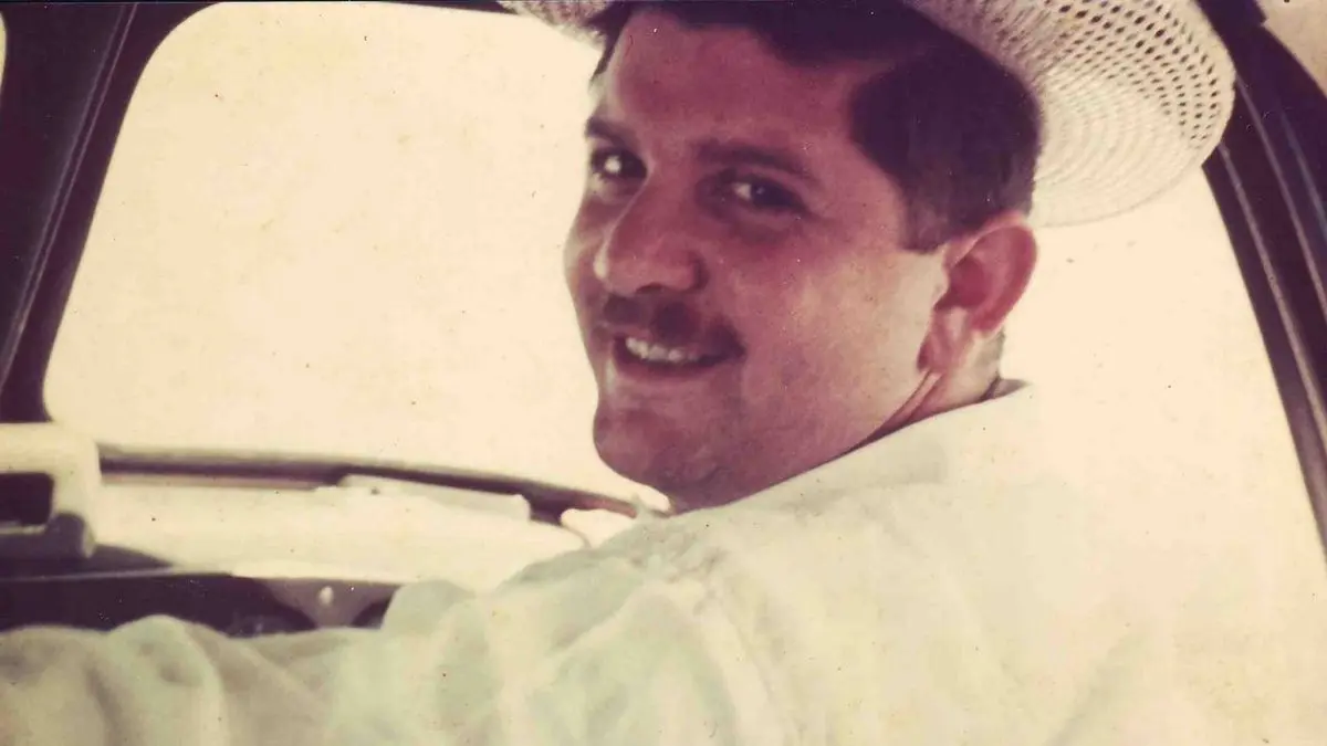 Homem sorridente em veículo, usando chapéu de palha e camisa branca.