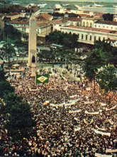 Manifestação pública com multidão, estátuas e edifícios públicos.