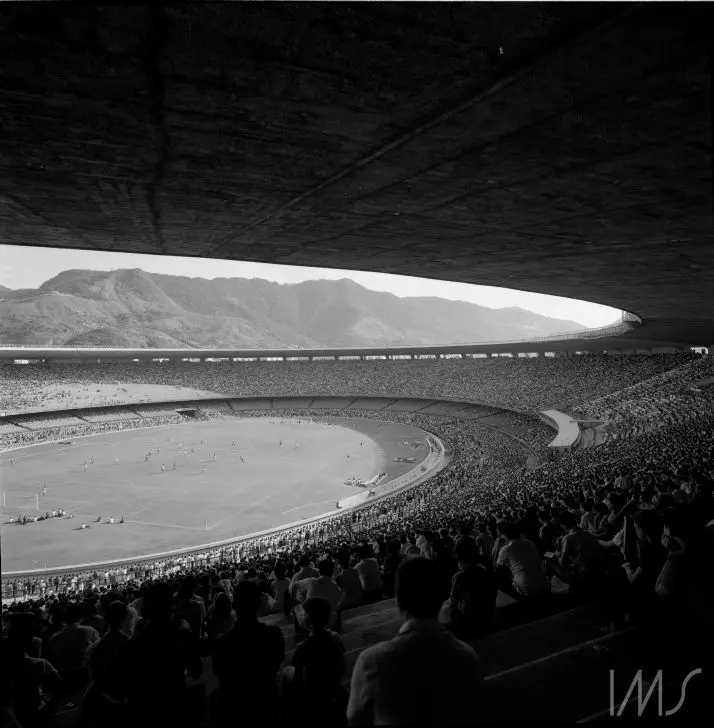 Estádio lotado durante jogo de futebol, com arquibancada cheia e montanhas ao fundo.