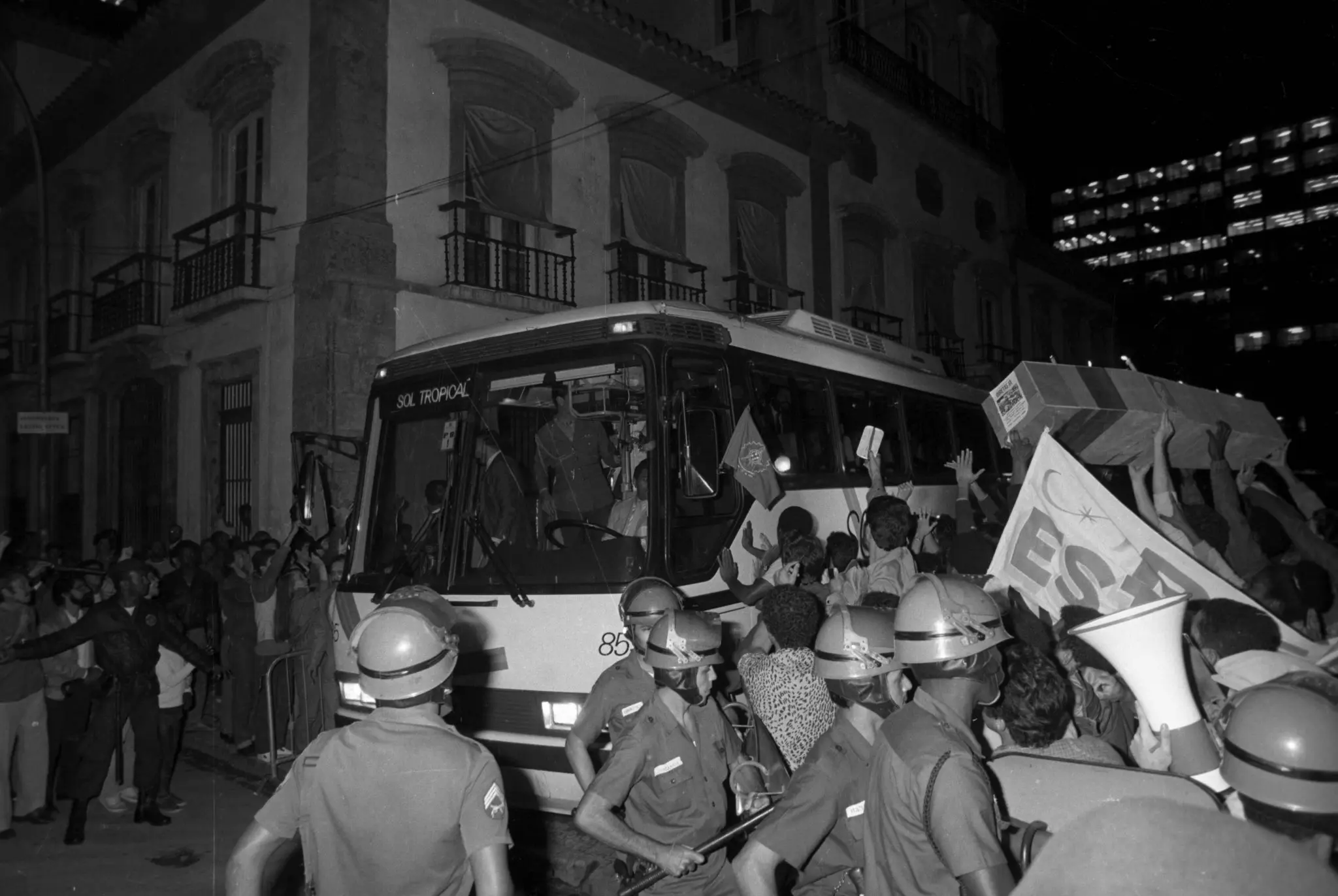 Fotografia preto e branco de pessoas aglomeradas ao redor de um ônibus "Sol Tropical" num cenário urbano misto de antigos e modernos prédios.