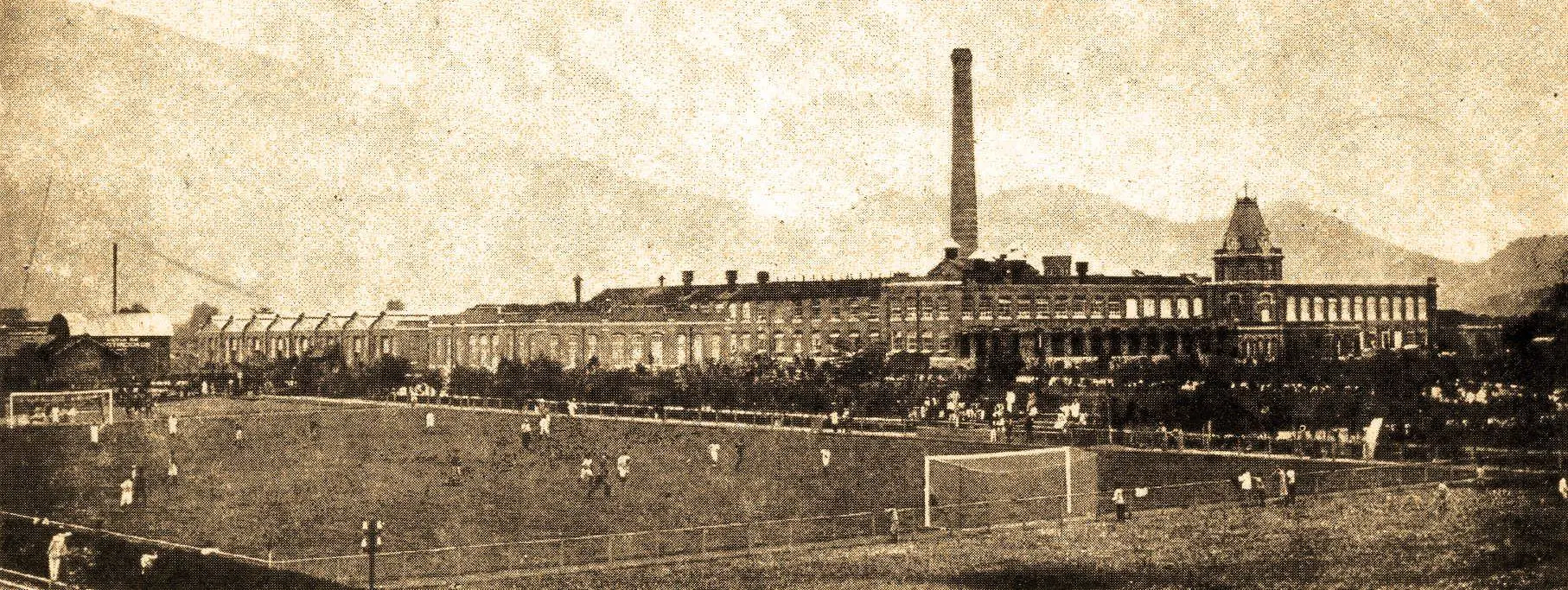 Fábrica e campo de futebol em preto e branco, com edifícios industriais e montanhas nebulosas ao fundo.