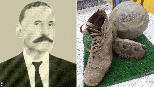Homem em terno clássico ao lado de chuteira e bola de futebol antiga.