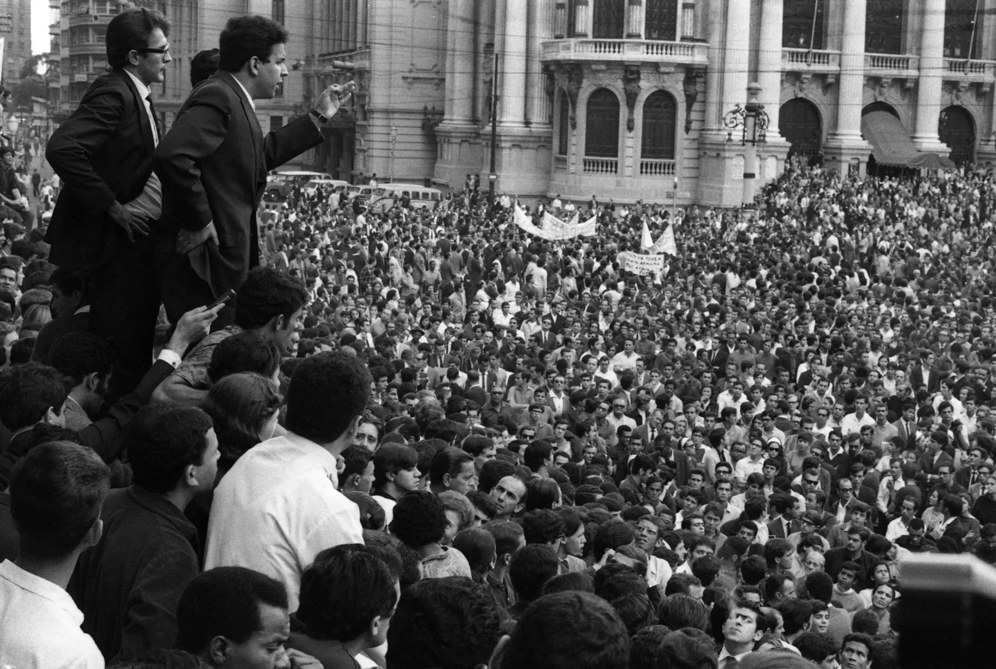 Manifestação em preto e branco com multidão密集 e homens em ternos discursando.