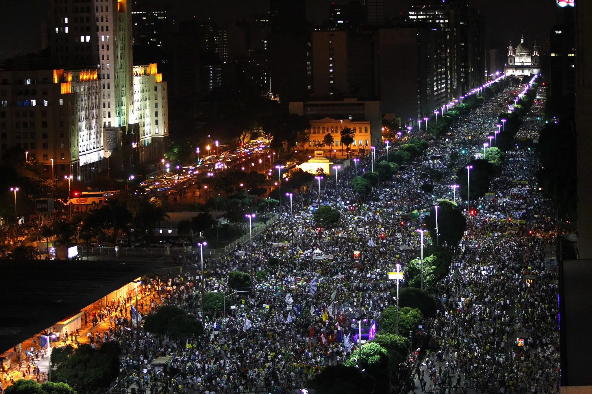 Manifestação noturna em avenida urbana com multidão e edifícios iluminados.