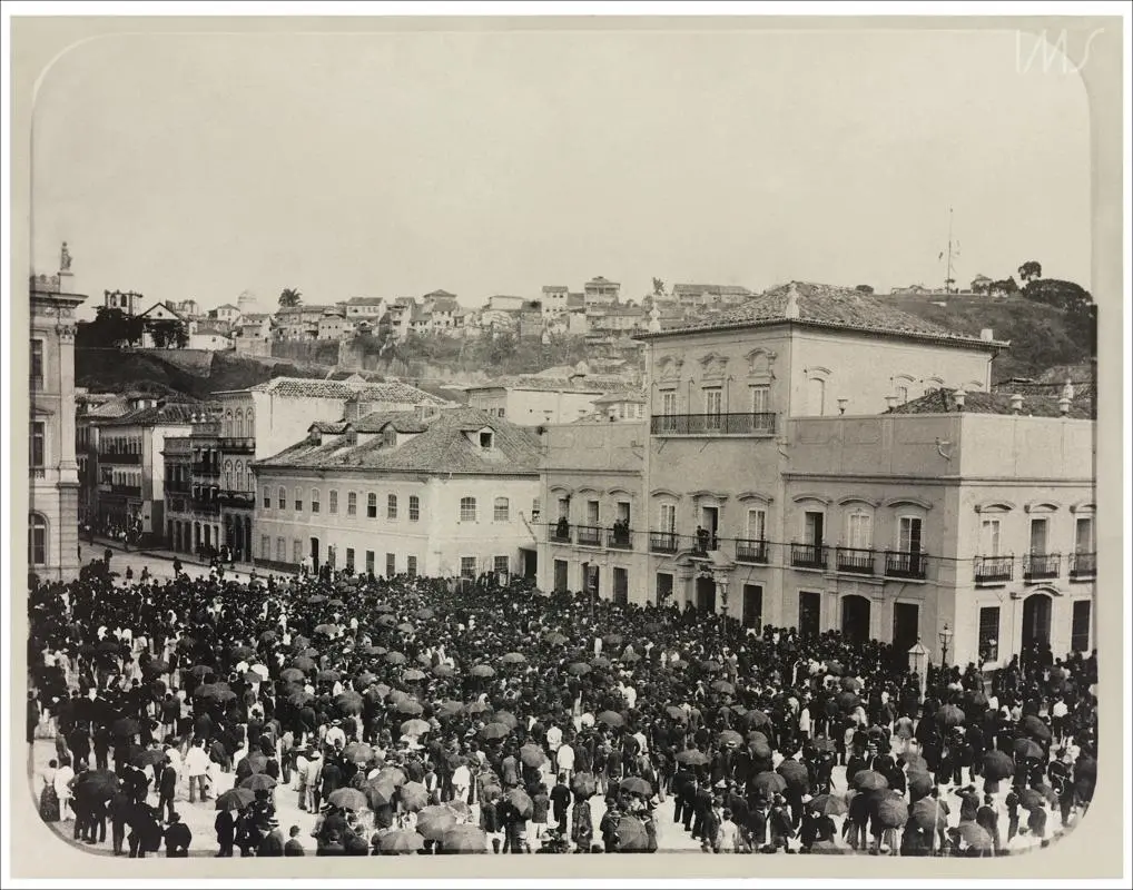 Multidão em praça histórica, com edifícios e colinas ao fundo.