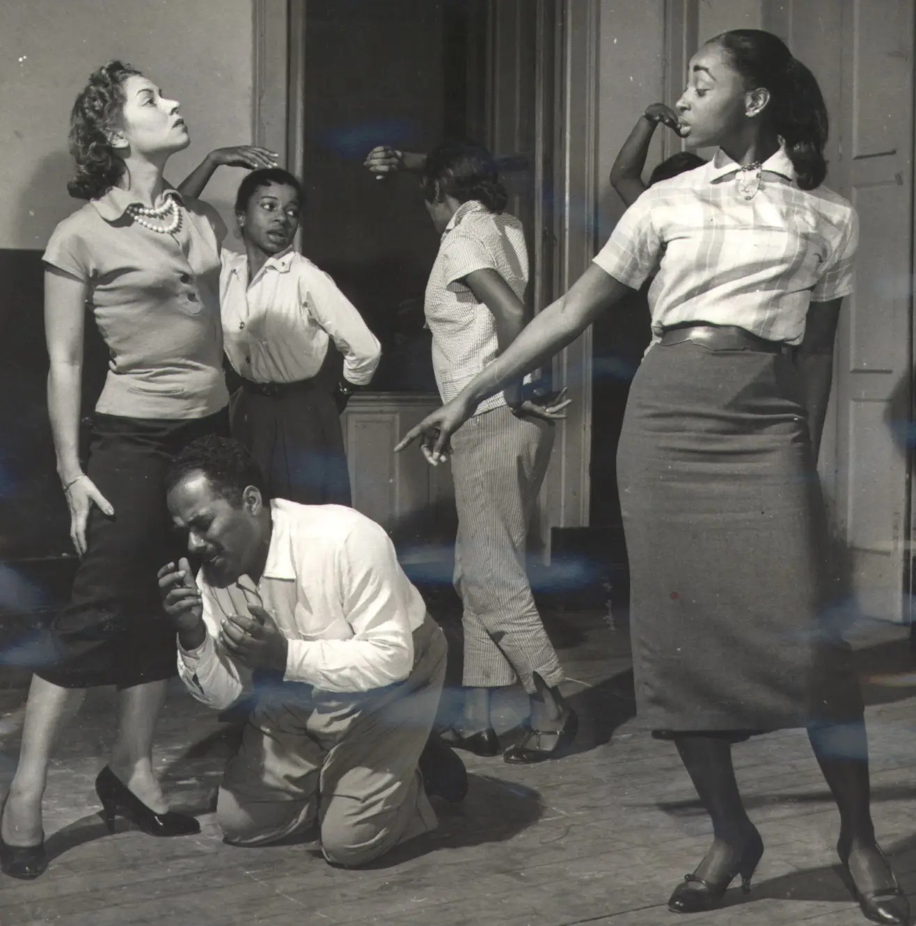 Seis pessoas em cenário teatral, duas mulheres de pé à esquerda, uma sentada no chão à direita.