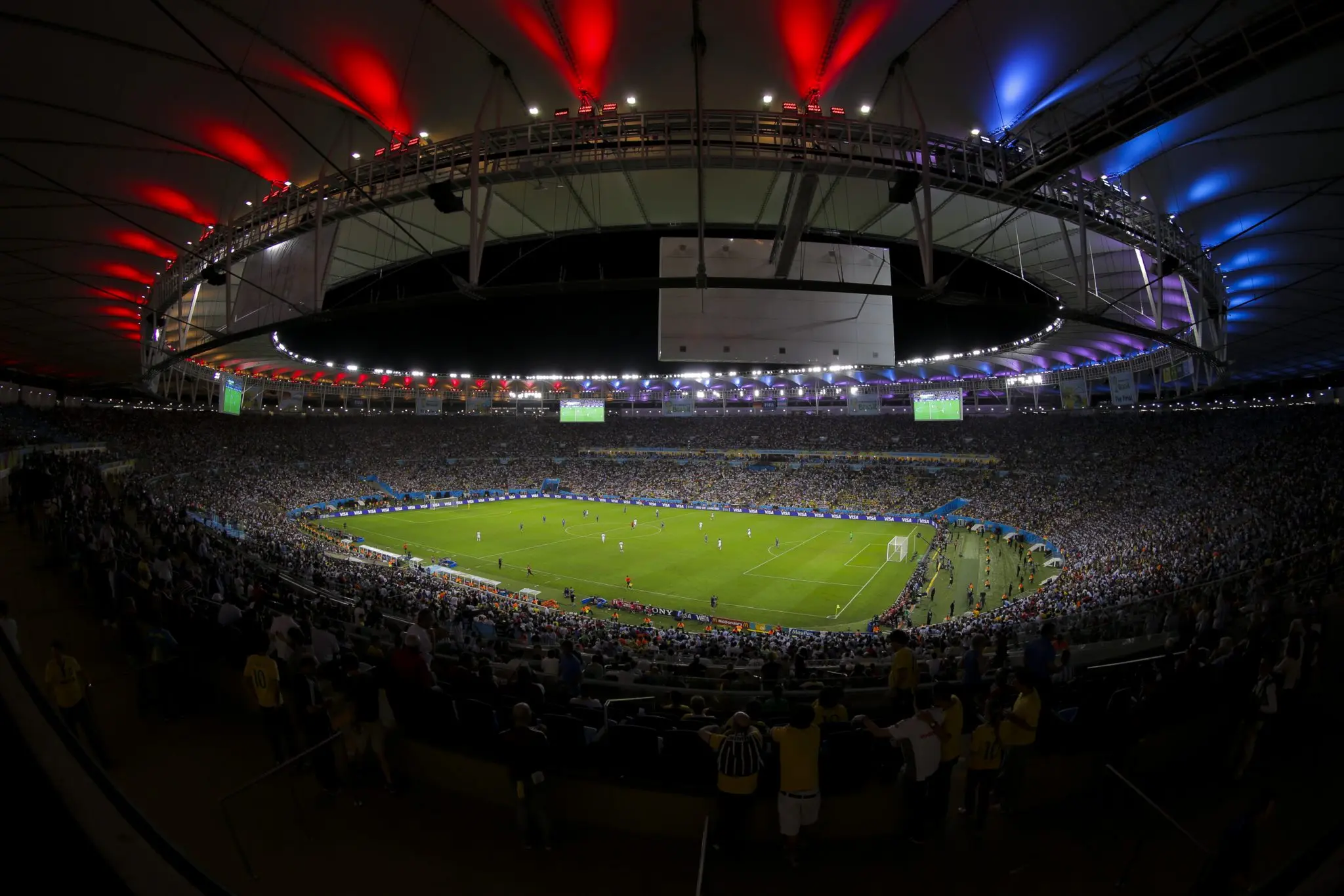 Estádio lotado durante partida de futebol com público e jogadores em ação.
