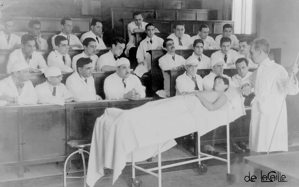 Aula médica em preto e branco com paciente em maca, médico explicando e estudantes assistindo.