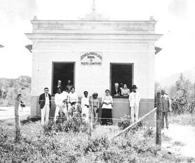 Grupo de pessoas em frente a Posto Sanitário, com edifício e vegetação ao fundo.