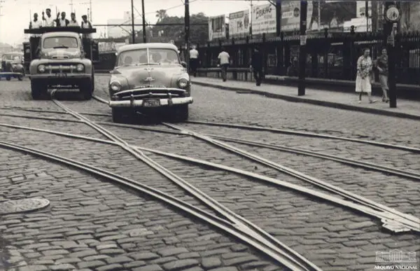 Rua com trilhos de bondes, carros antigos e edifícios publicitários.