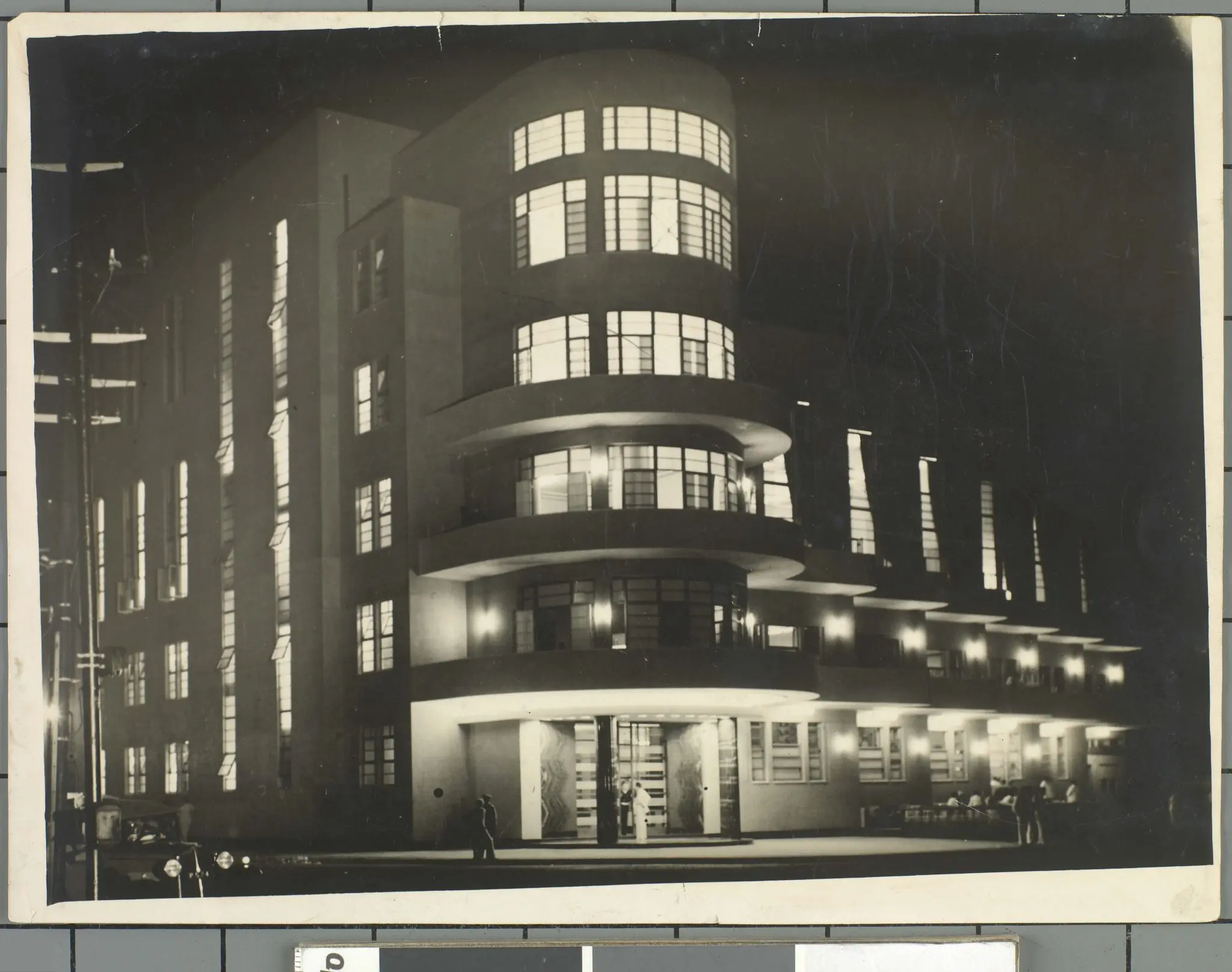 Edifício modernista à noite, construções distintas e pessoas caminhando.