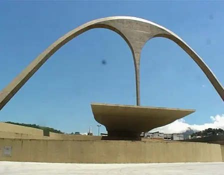 Monumento modernista com estrutura curvada em concreto e céu azul ao fundo.